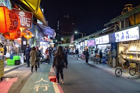 A bustling street market at night with vibrant lanterns hanging above. The market is lined with food stalls and small shops, each illuminated with bright signage. People are walking along the street, some stopping to purchase goods or wait in line at the stalls. The atmosphere is lively and inviting.