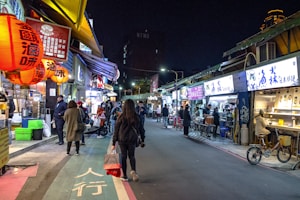 A bustling street market at night with vibrant lanterns hanging above. The market is lined with food stalls and small shops, each illuminated with bright signage. People are walking along the street, some stopping to purchase goods or wait in line at the stalls. The atmosphere is lively and inviting.