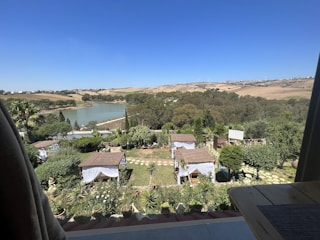 A serene landscape featuring a view from a balcony overlooking a garden with small gazebos covered by tile roofs. The garden is lush with trees, shrubs, and multiple potted plants. Beyond the garden, there is a large lake surrounded by dense greenery and further in the distance, rolling hills under a clear blue sky.