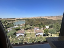 A serene landscape featuring a view from a balcony overlooking a garden with small gazebos covered by tile roofs. The garden is lush with trees, shrubs, and multiple potted plants. Beyond the garden, there is a large lake surrounded by dense greenery and further in the distance, rolling hills under a clear blue sky.