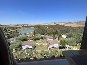 A serene landscape featuring a view from a balcony overlooking a garden with small gazebos covered by tile roofs. The garden is lush with trees, shrubs, and multiple potted plants. Beyond the garden, there is a large lake surrounded by dense greenery and further in the distance, rolling hills under a clear blue sky.