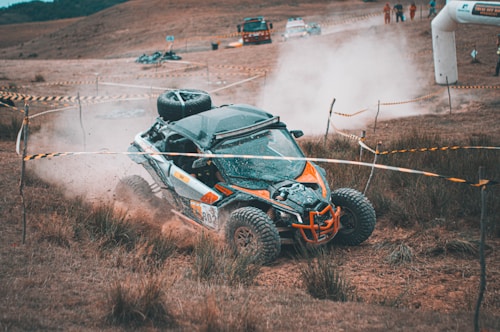 A rugged off-road vehicle navigates a dirt track, surrounded by brown grass and marked off by yellow caution tape. The vehicle kicks up a cloud of dust as it maneuvers through the terrain, while spectators and other vehicles are visible in the background.