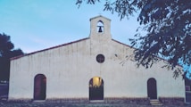 A rustic, white stucco church with a simple architectural design features three arched doorways and a small belfry on top. The church is surrounded by a serene outdoor setting with trees, and the sky is a light blue, suggesting early evening or morning.
