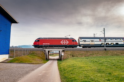 A modern passenger train in red and white colors travels on a raised railway track above a small road. The sky is overcast, and the surrounding landscape includes green grass and a blue building on the left side.