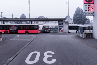 A road with the number 30 painted on it leads to a small bus station. A red bus is parked on the left, displaying an advertisement. In the background, a train passes behind the station structure, and a white bus is parked on the right. A sign with fuel prices is visible to the right, and trees and a building are in the distance.