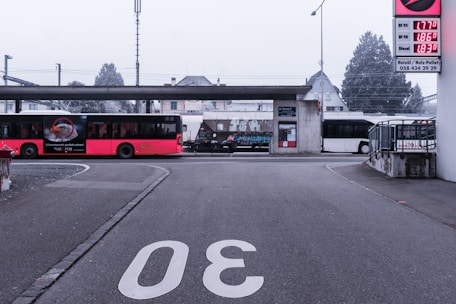 A road with the number 30 painted on it leads to a small bus station. A red bus is parked on the left, displaying an advertisement. In the background, a train passes behind the station structure, and a white bus is parked on the right. A sign with fuel prices is visible to the right, and trees and a building are in the distance.