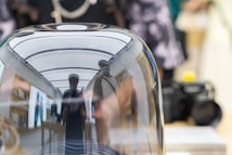 A reflective surface prominently features distorted reflections of several people and objects. The background of the reflection shows a brightly lit retail environment with tech products displayed. There are elements such as a camera visible in the periphery.