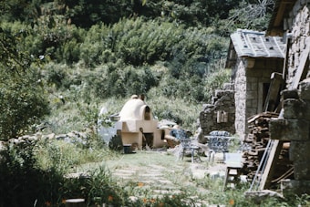 A rustic outdoor scene features a stone building with a tiled roof on the right, surrounded by lush greenery. A round, clay oven or kiln sits in the center of the image, with metal chairs and a table nearby. Wooden logs and other materials are piled up, suggesting a rural or garden setting. The area is bathed in sunlight, enhancing the natural and serene atmosphere.