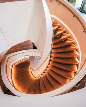 A spiral staircase with wooden steps and curved white railings illuminated by soft lighting embedded along the edges. The design is modern and elegant, creating a sense of fluidity and motion as it spirals downward.