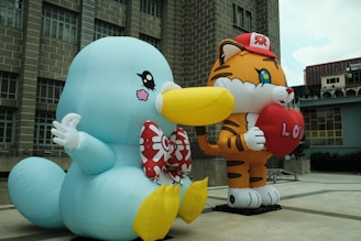 Two large inflatable cartoon characters are prominently displayed outdoors. One character resembles a blue bird-like creature with a large yellow beak and a red bow tie, while the other character is a tiger wearing a red cap and holding a red heart with 'LOVE' written on it. The background consists of a building with grid-like windows.