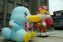 Two large inflatable cartoon characters are prominently displayed outdoors. One character resembles a blue bird-like creature with a large yellow beak and a red bow tie, while the other character is a tiger wearing a red cap and holding a red heart with 'LOVE' written on it. The background consists of a building with grid-like windows.