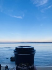 A Ghirardelli-branded coffee cup is held in the foreground against a backdrop of a tranquil sea and a clear blue sky. The horizon blends seamlessly with the calm water, while a few figures stand on the sandy beach.