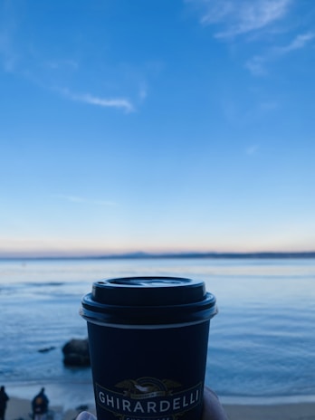 A Ghirardelli-branded coffee cup is held in the foreground against a backdrop of a tranquil sea and a clear blue sky. The horizon blends seamlessly with the calm water, while a few figures stand on the sandy beach.