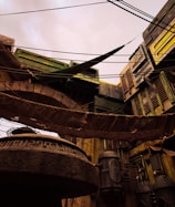 A futuristic cityscape with industrial architecture featuring large pipes and angular structures. There are cables and fabric draped across the scene, and the buildings have a worn, weathered appearance.
