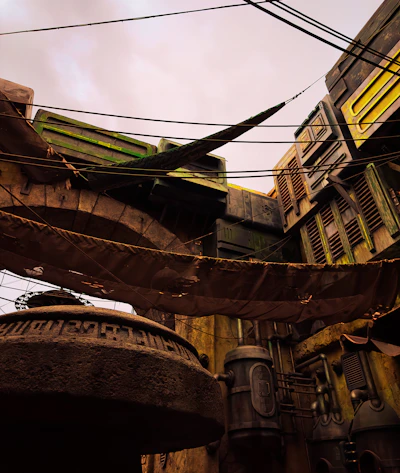 A futuristic cityscape with industrial architecture featuring large pipes and angular structures. There are cables and fabric draped across the scene, and the buildings have a worn, weathered appearance.