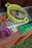 A yellow plastic container filled with sand, leaves, and a small purple toy shovel is positioned on a patterned surface. Nearby, there is a purple cup with sand and a green mold, possibly for playing with sand. The setting appears to be a wooden surface outdoors, possibly a play area.