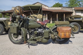 A vintage military motorcycle with olive green paint is parked on a gravel surface. It features a leather saddle seat and a brown leather side bag attached to the rear. Nearby, there are similarly colored military vehicles with visible stars and serial numbers. A camouflage helmet is placed on the motorcycle handlebar, and a rustic building with stone walls and a wooden porch is in the background, along with green foliage.