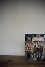 A detailed and ornate album cover featuring a collage of intricate artwork with a central focus on eyes peering through a decorative mask. Surrounding elements include various figures and animals intertwined with baroque-style gold elements, against a dark background. The cover appears on a wooden surface with a plain wall backdrop.