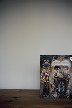 A detailed and ornate album cover featuring a collage of intricate artwork with a central focus on eyes peering through a decorative mask. Surrounding elements include various figures and animals intertwined with baroque-style gold elements, against a dark background. The cover appears on a wooden surface with a plain wall backdrop.