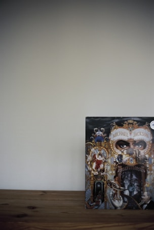 A detailed and ornate album cover featuring a collage of intricate artwork with a central focus on eyes peering through a decorative mask. Surrounding elements include various figures and animals intertwined with baroque-style gold elements, against a dark background. The cover appears on a wooden surface with a plain wall backdrop.