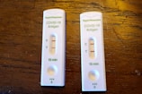 Two COVID-19 antigen rapid test kits are placed on a wooden surface. Each kit displays two lines on the result window indicating positive results. The tests are labeled with 'Rapid Response' along with 'COVID-19 Antigen' and show that results are available in 15 minutes.