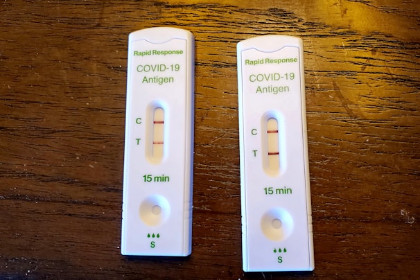 Two COVID-19 antigen rapid test kits are placed on a wooden surface. Each kit displays two lines on the result window indicating positive results. The tests are labeled with 'Rapid Response' along with 'COVID-19 Antigen' and show that results are available in 15 minutes.