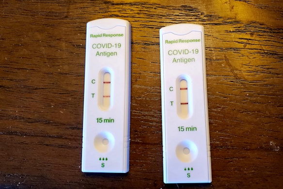 Two COVID-19 antigen rapid test kits are placed on a wooden surface. Each kit displays two lines on the result window indicating positive results. The tests are labeled with 'Rapid Response' along with 'COVID-19 Antigen' and show that results are available in 15 minutes.