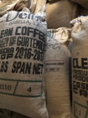 Several burlap sacks labeled as clean coffee, originating from Guatemala, are stacked together. The sacks display text including the year 2016-2017 and other product details. The texture of the sacks is coarse and natural.