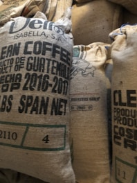 Several burlap sacks labeled as clean coffee, originating from Guatemala, are stacked together. The sacks display text including the year 2016-2017 and other product details. The texture of the sacks is coarse and natural.