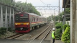 A train is approaching on a railway track surrounded by lush greenery and infrastructure. A person wearing a neon safety vest and helmet is observing the train near the tracks. Overhead electric lines and poles are visible as well.