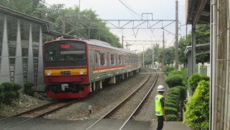 A train is approaching on a railway track surrounded by lush greenery and infrastructure. A person wearing a neon safety vest and helmet is observing the train near the tracks. Overhead electric lines and poles are visible as well.