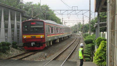 A train is approaching on a railway track surrounded by lush greenery and infrastructure. A person wearing a neon safety vest and helmet is observing the train near the tracks. Overhead electric lines and poles are visible as well.
