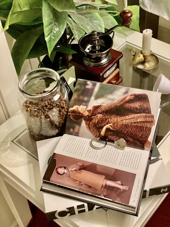 A stylish setup on a glass table includes a book opened to a fashion magazine spread showing models in vintage clothing. Beside it, a decorative magnifying glass rests on the pages. A jar filled with coffee beans and a vintage manual coffee grinder are adjacent to a candle on a brass holder. In the background, lush green leafy plants provide a natural touch.