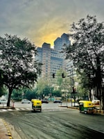 A bustling city intersection with a prominent modern high-rise building in the background, framed by large trees on either side. The road is busy with autorickshaws and cars, and traffic signals are visible. The sky is overcast with a soft, yellowish glow.