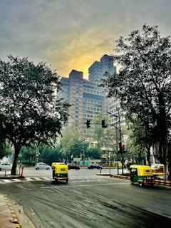 A bustling city intersection with a prominent modern high-rise building in the background, framed by large trees on either side. The road is busy with autorickshaws and cars, and traffic signals are visible. The sky is overcast with a soft, yellowish glow.
