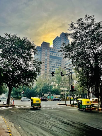 A bustling city intersection with a prominent modern high-rise building in the background, framed by large trees on either side. The road is busy with autorickshaws and cars, and traffic signals are visible. The sky is overcast with a soft, yellowish glow.