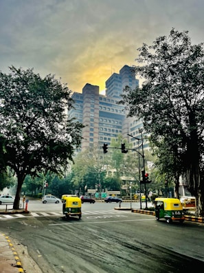A bustling city intersection with a prominent modern high-rise building in the background, framed by large trees on either side. The road is busy with autorickshaws and cars, and traffic signals are visible. The sky is overcast with a soft, yellowish glow.