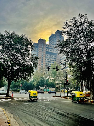 A bustling city intersection with a prominent modern high-rise building in the background, framed by large trees on either side. The road is busy with autorickshaws and cars, and traffic signals are visible. The sky is overcast with a soft, yellowish glow.