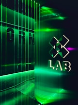 A futuristic interior with green LED lighting casting a vibrant glow. A geometric logo with the word 'LAB' is visible on a reflective wall, alongside vertical light strips creating an immersive atmosphere.