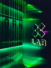 A futuristic interior with green LED lighting casting a vibrant glow. A geometric logo with the word 'LAB' is visible on a reflective wall, alongside vertical light strips creating an immersive atmosphere.