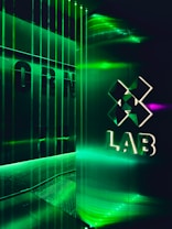 A futuristic interior with green LED lighting casting a vibrant glow. A geometric logo with the word 'LAB' is visible on a reflective wall, alongside vertical light strips creating an immersive atmosphere.