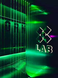 A futuristic interior with green LED lighting casting a vibrant glow. A geometric logo with the word 'LAB' is visible on a reflective wall, alongside vertical light strips creating an immersive atmosphere.