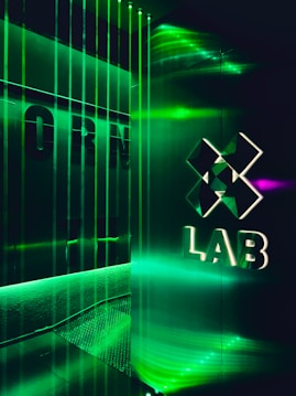 A futuristic interior with green LED lighting casting a vibrant glow. A geometric logo with the word 'LAB' is visible on a reflective wall, alongside vertical light strips creating an immersive atmosphere.