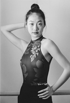 A black and white portrait of a woman posing gracefully with one hand on her hip and the other behind her head. She is wearing a floral-patterned sleeveless top, her hair is styled in a neat bun, and she gazes confidently at the camera.