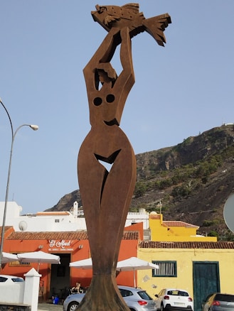 A tall, abstract metal sculpture of a human figure holding a fish above its head is prominently displayed in an outdoor area. The sculpture features stylized shapes and cutouts, standing against a backdrop of colorful buildings with red and yellow walls. In the distance, a rocky hill with sparse vegetation is visible under a clear blue sky.