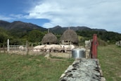 A rural farm setting with several pigs gathered in a pen in front of two thatched-roof structures. The backdrop features rolling hills and lush greenery under a partly cloudy sky. A stone wall and metal gate are in the foreground, with a metal container nearby.