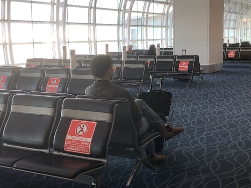 A person wearing a mask sits in an airport seating area with social distancing signs on many empty seats. The area is spacious, with large windows allowing natural light in, and there are only a few people scattered around.