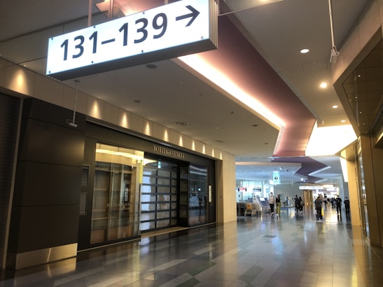 A modern indoor airport corridor with polished flooring. Above, a large illuminated sign indicates gates 131-139. On the left, a closed high-end store with a dark facade and signage that reads 'Bottega Veneta'. Farther down the corridor, a few travelers are visible, some pushing luggage carts under the well-lit ceiling with recessed lighting.