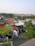 A residential area with red-tiled rooftops, green trees, and a few buildings. The view extends to the horizon showing a mix of vegetation and urban structures. The sky is clear and houses are closely packed, indicating a densely populated area.