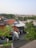 A residential area with red-tiled rooftops, green trees, and a few buildings. The view extends to the horizon showing a mix of vegetation and urban structures. The sky is clear and houses are closely packed, indicating a densely populated area.